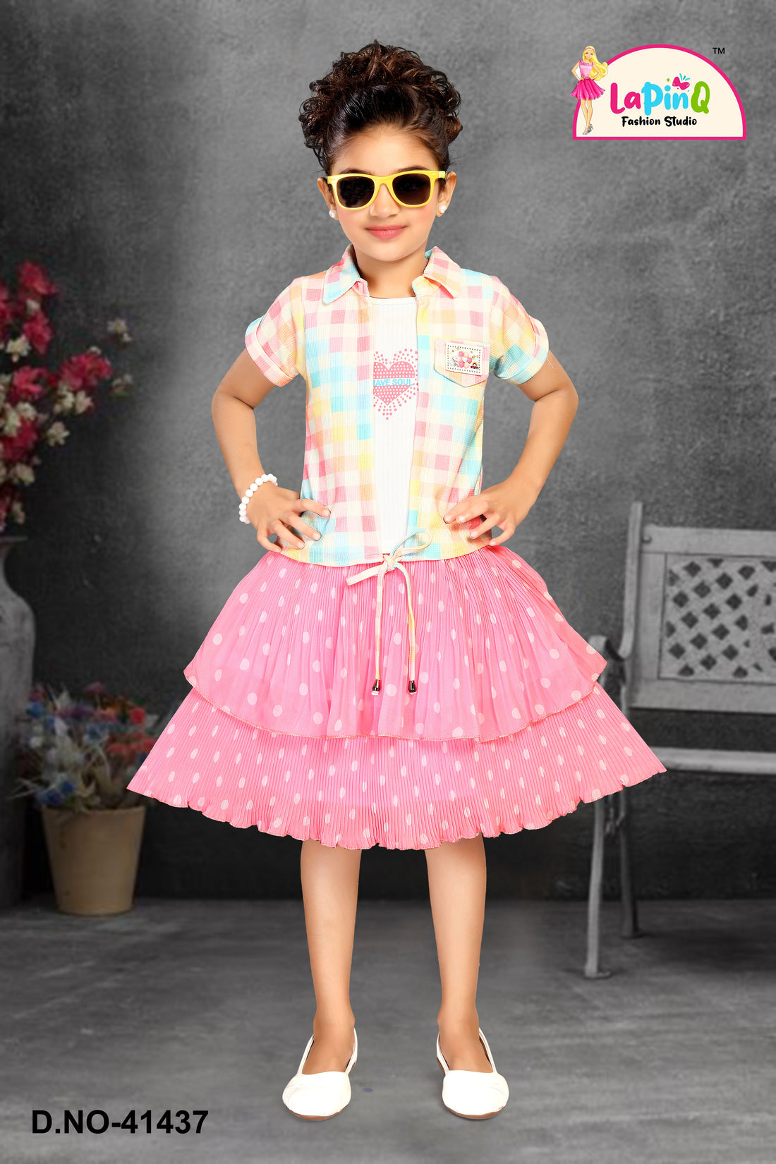 Young girl in a colorful checkered shirt and pink skirt with polka dots, wearing sunglasses, against a gray background.
