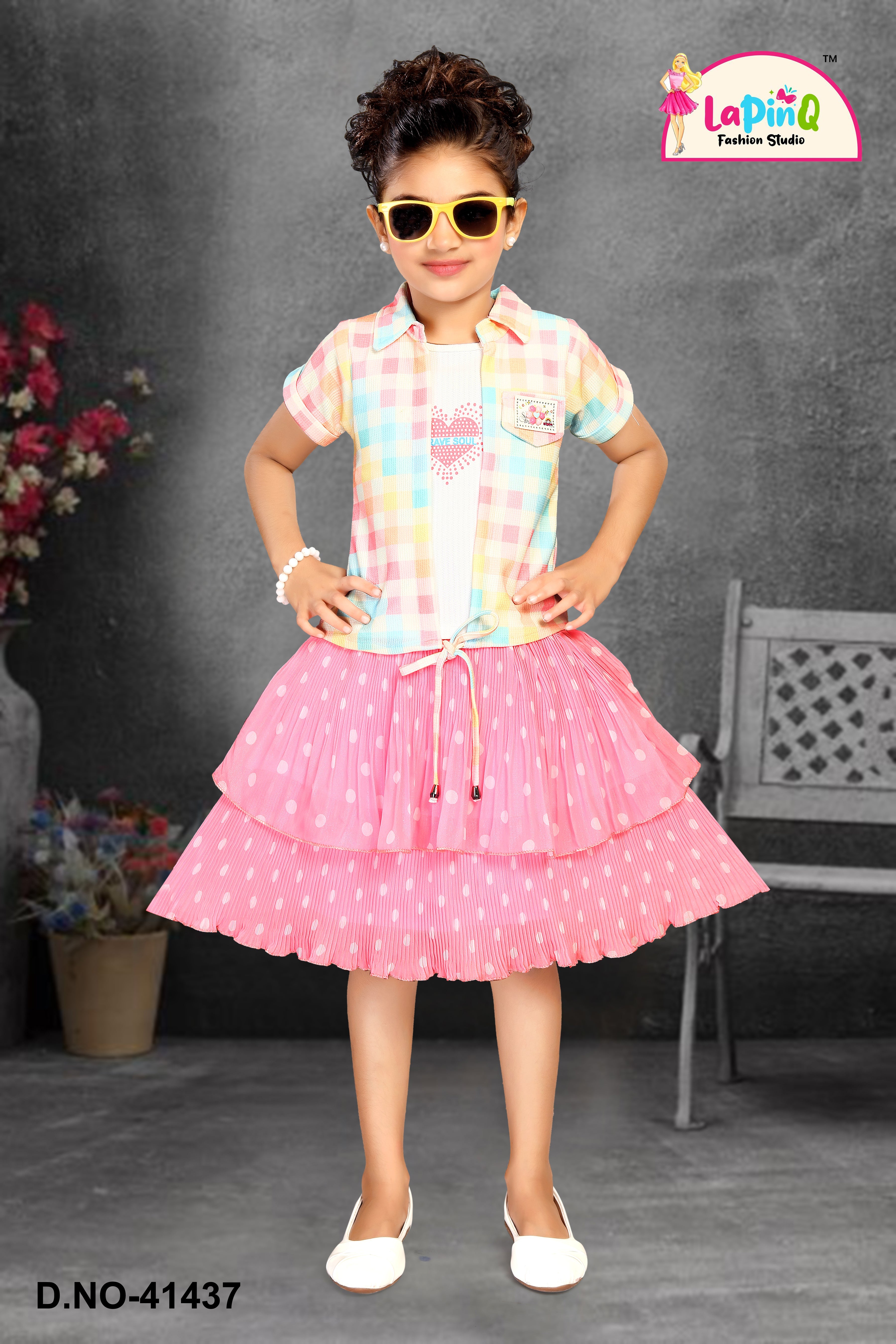 Young girl in a colorful checkered shirt and pink skirt with polka dots, wearing sunglasses, against a gray background.